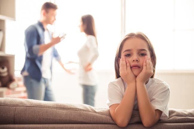 Sad little girl looks straight ahead while her parents are arguing in the background.
