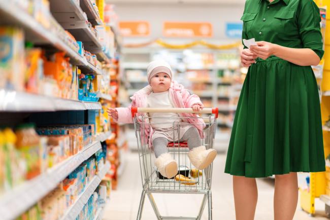 A mother and her baby sitting in a grocery cart choosing food in a supermarket aisle.