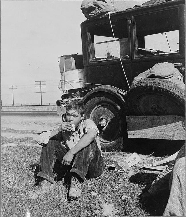 A boy eating a sandwich near a packed car on the grass by the side of a road.