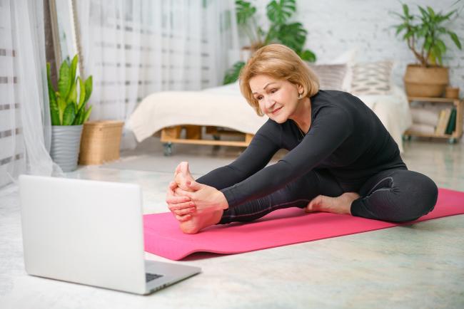 Older woman sitting on yoga mat stretches as part of online fitness training.