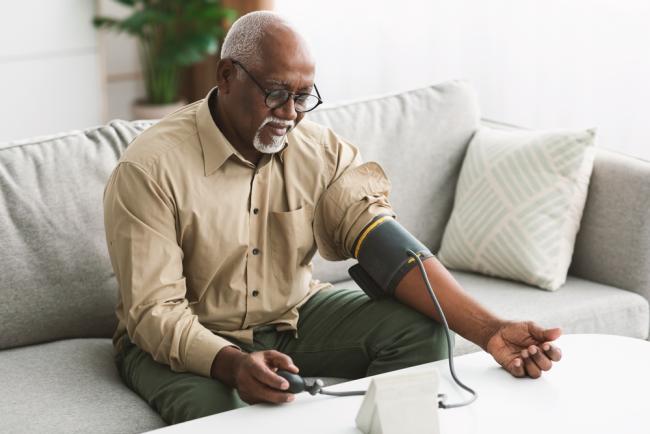 Black male measuring blood pressure sitting on a couch at home.