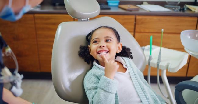 Young child smiles, pointing to her teeth, during dental checkup.