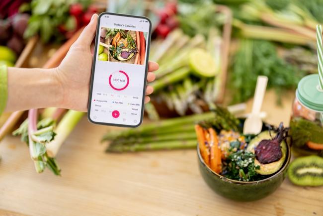 Holding smart phone with running mobile application for track calories consumed on background of healthy food ingredients. Concept of modern technology in proper nutrition
