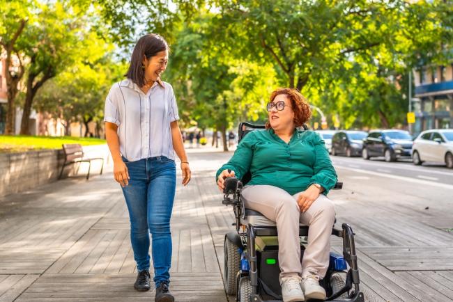 Woman using an electric wheelchair and talking to a friend walking alongside her on a city street