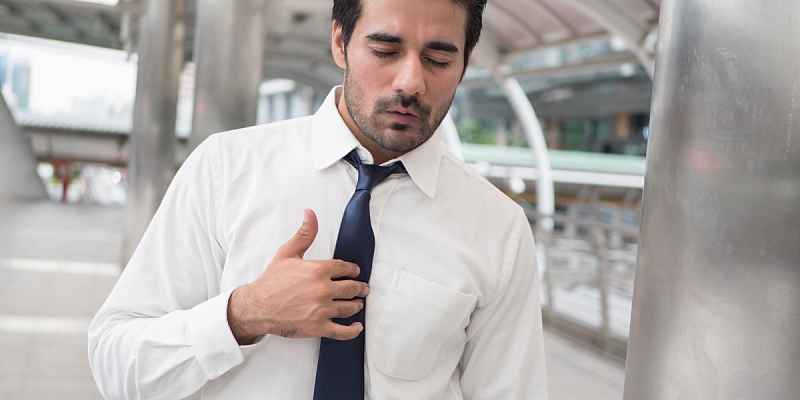 A man in a white shirt and tie, indoors, pressing down on his chest with an uncomfortable expression, experiencing heartburn.