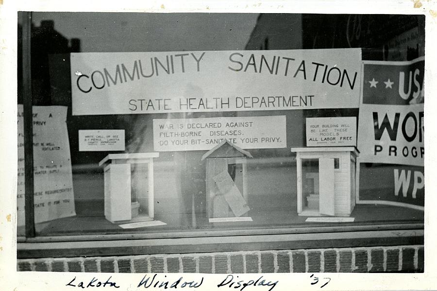 Exterior view of window display showing three model privies under a sign that reads “Community Sanitation”