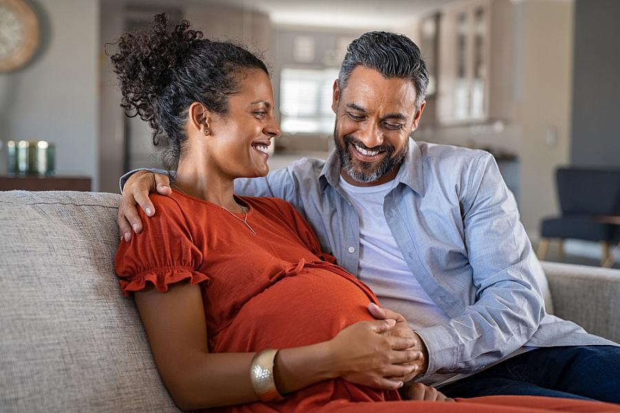 Pregnant woman on couch with man, both smiling and touching her belly.