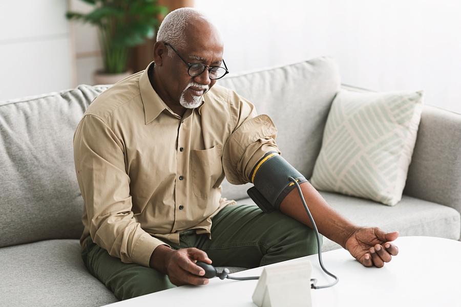 Black male measuring blood pressure sitting on a couch at home.