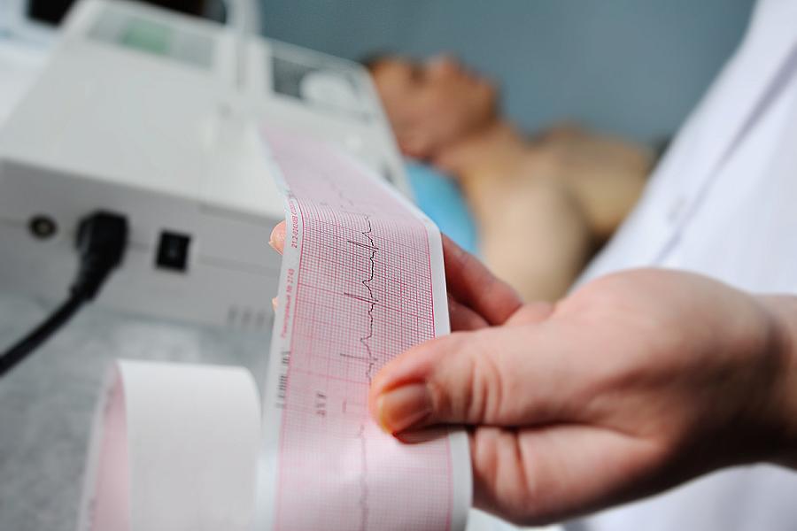 Hand holding ECG printout with man lying in bed in background.