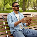 A vision impaired man sitting on a park bench and reading a braille book