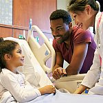 A young girl in a hospital bed smiling at her father and pediatrician