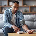 Photo of young African-American man sitting on a couch and checking his own blood pressure with upper arm cuff and monitor.  