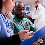 Close up of african american man reviewing medical records along with asian female member of in house hospital staff. Doctor indicating treatment to young male patient at sanatorium lobby.