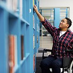 Student in wheelchair choosing books from shelf in library.