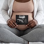 Unrecognizable black pregnant lady demonstrating her baby sonography photo while sitting on bed at home, showing firts photo of her child, enjoying happy maternity time, cropped image, closeup