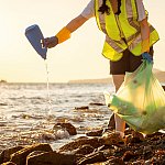 Person in a safety vest cleaning up debris from the ocean and a rocky shore. They are pouring water out of a plastic, liquid-dispensing bottle that was collected. 