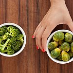 A woman’s hand holding a bowl of brussel sprouts, with a bowl of cooked broccoli to the left.