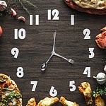 Black wooden clock with utensils as hands and surrounded by food