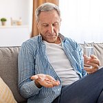 Older man sitting on a couch with a glass of water, looking at pills in his hand.