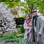 Senior man with a walker and a younger woman strolling outdoors in an urban park.