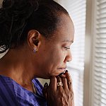 An older black woman mournfully looks out a window.