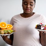 Black woman holding plates with one plate with fruits and one with cakes and donuts, looking at the unhealthy plate with skepticism.