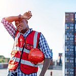 Black male engineer in uniform on a sunny day holding a hard hat and wiping sweat from his brown.