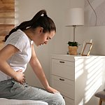 Young woman sitting on bed and clutching her stomach in pain.