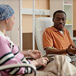 A black man and a white woman with a head scarf talking during chemotherapy treatment in clinic.