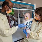 Two doctors, wearing masks, interact with a smiling infant patient. 