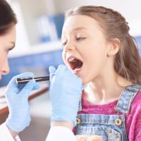 Woman doctor examining girl’s throat with spatula and flashlight