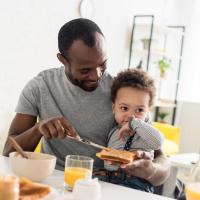 Father with young son on his lap spreading peanut butter on toast.