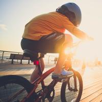 A young boy riding his bicycle on the boardwalk by the beach backlit by the sun wearing a helmet.