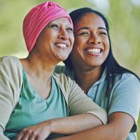A caregiver and a cancer patient wearing a headscarf sitting close together and smiling.