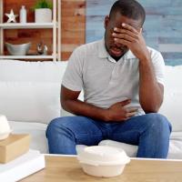 A man sitting on a couch holding his stomach in pain, with takeout food containers on a table in front of him.