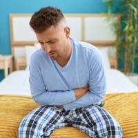 A man sitting at the edge of his bed, holding his stomach in pain.