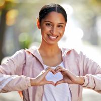 A smiling woman outdoors making a heart shape with her hands.
