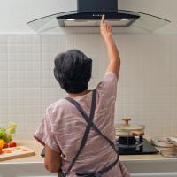 A woman cooking in a kitchen, reaching up to adjust the range hood above the stove.