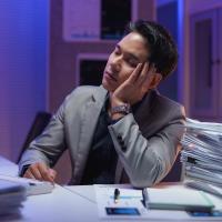 Person dozing off at workplace with head in hand, surrounded by paperwork and computer in a dimly lit office