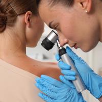 Doctor examining young woman's mole with dermatoscope in hospital, closeup Graphic