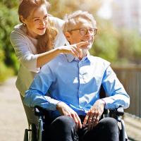 A female caregiver pushing an older man in a wheelchair outdoors.