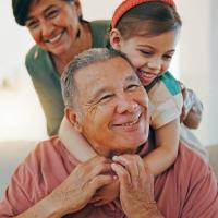 An older man smiling while a child hugs him from behind, with an older woman nearby.