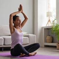 A woman sitting on a yoga mat indoors with her hands raised above her head.