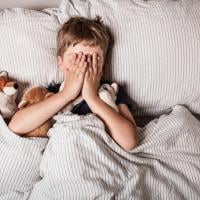 A young boy covers his face with his hands at bedtime. 