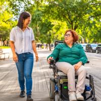 Woman using an electric wheelchair and talking to a friend walking alongside her on a city street