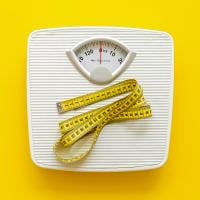 A bathroom scale with a measuring tape draped across it on a bright yellow background.