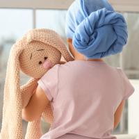 A child wearing a headscarf looking toward a window while holding a stuffed animal.
