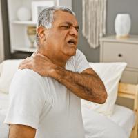 An older man seated on a bed holding his shoulder in pain.