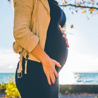 A pregnant woman standing beside the water with her hands resting on her belly.