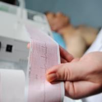 Hand holding ECG printout with man lying in bed in background.